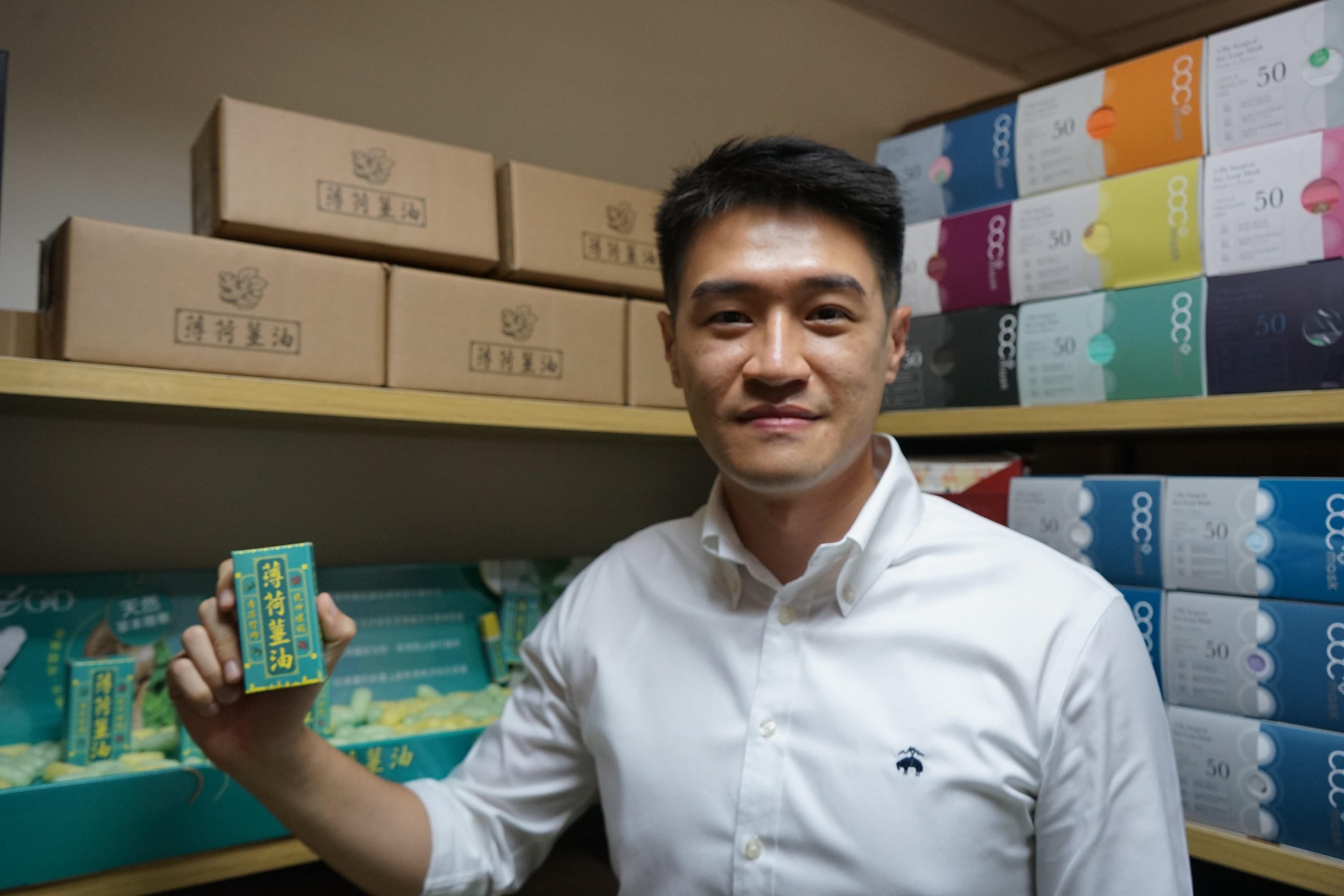 身穿白色上衣的 Dennis 手持绿色 G&D 产品盒，身后的层架摆放 acc+ 口罩